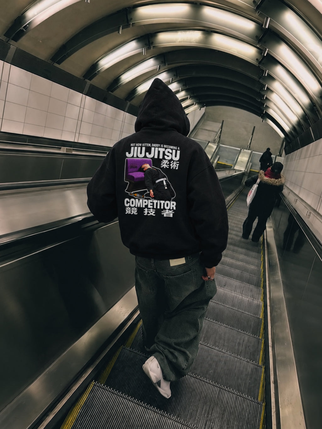 Black Not Now Kitten hoodie from BJJ World, back print worn hood up on a subway escalator.