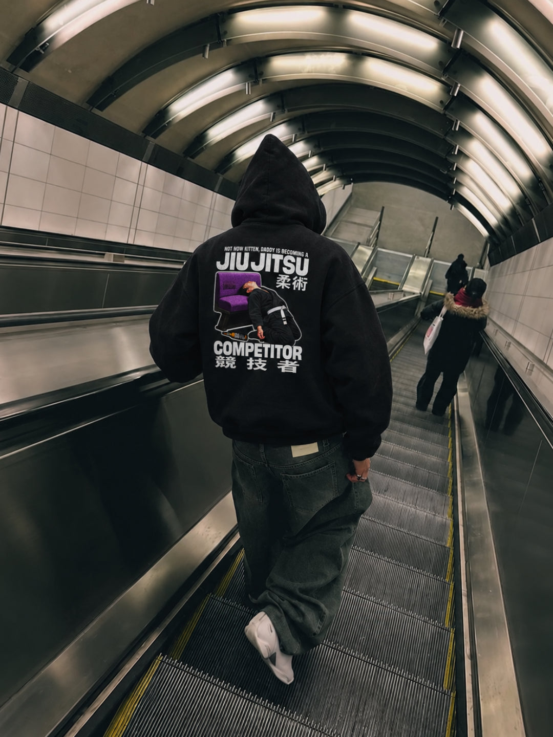 Black Not Now Kitten hoodie from BJJ World, back print worn hood up on a subway escalator.