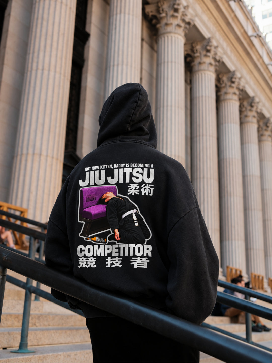 Black Not Now Kitten hoodie from BJJ World, back print worn hood up on stone steps.