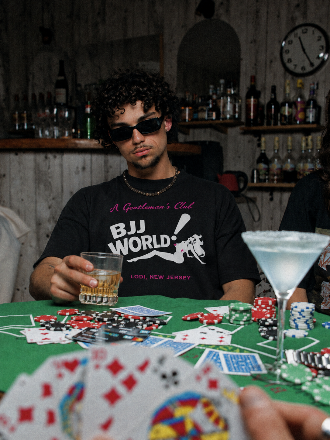 Black A Gentleman's Club tee from BJJ World, pink and white front print worn at a card table.