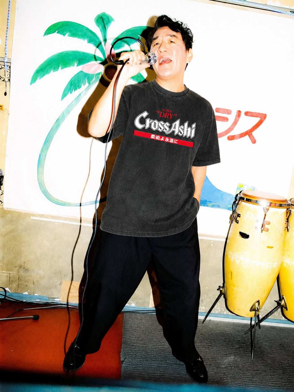 Washed black CrossAshi tee from BJJ World, worn at a microphone beside congas.